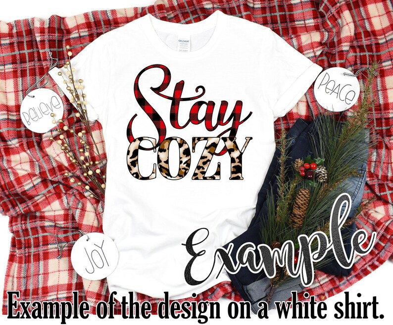 May include: White t-shirt with the words "Stay Cozy" in a mix of red plaid and leopard print. The shirt is displayed on a red and white plaid blanket with decorative ornaments and a pair of blue jeans. The text "Example of the design on a white shirt" is also visible.
