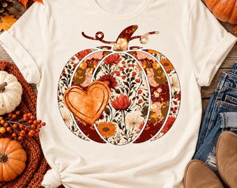 May include: A cream-colored short-sleeve t-shirt featuring a large central graphic of a pumpkin. The pumpkin is filled with a floral pattern in shades of orange, red, and brown, and includes a prominent orange heart shape on its left side.