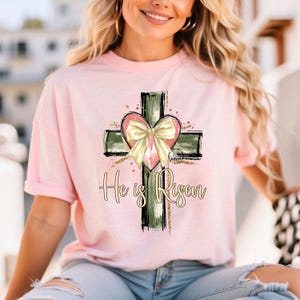 May include: A pink t-shirt with a green and black cross with a pink heart and a yellow bow. The text "He is Risen" is printed below the cross.
