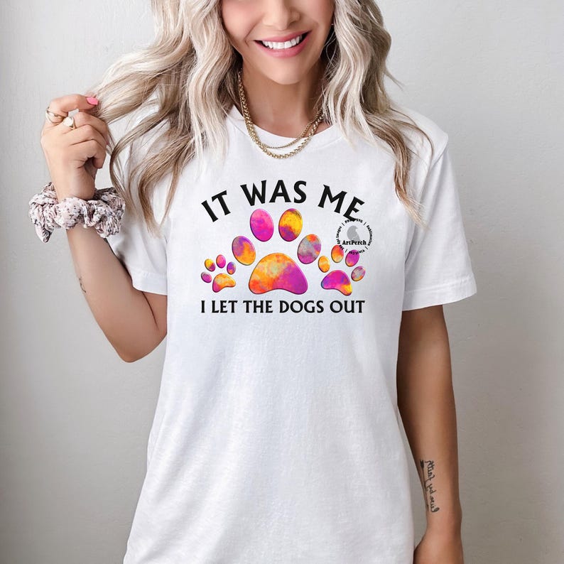 May include: White t-shirt with a colorful paw print design and the text "It was me I let the dogs out".