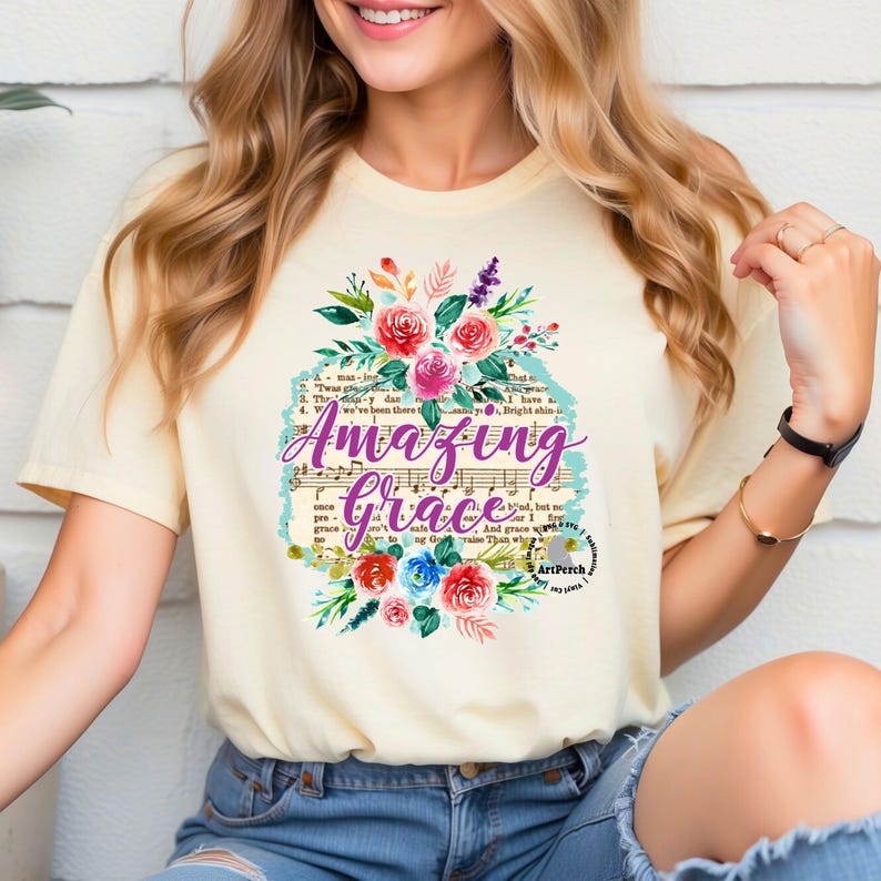 May include: A light yellow t-shirt with a floral design and the words "Amazing Grace" printed on it. The design includes musical notes and a vintage-style font.