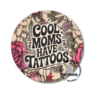 May include: Round sticker with the text "COOL MOMS HAVE TATTOOS" in black, surrounded by a floral design with pink roses and other flowers. The background is a beige color.