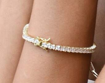Puede incluir: Un brazalete de tenis dorado con una fila de piedras cuadradas transparentes.