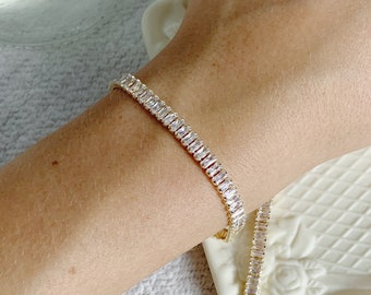 Puede incluir: Un collar y una pulsera de cadena dorada con piedras transparentes en forma de baguette. Las joyas están sobre una superficie blanca.