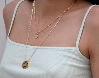 May include: A woman wearing a light green tank top with three gold necklaces. The bottom necklace has a round pendant with a design.