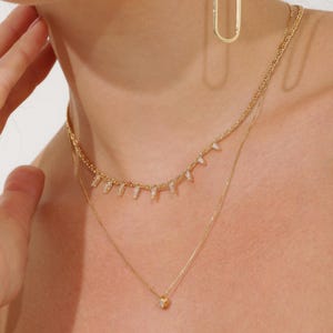 May include: A layered gold necklace set. The top necklace features small, clear, rectangular stones. The second necklace is a delicate chain with a small, round pendant. A gold hoop earring is visible in the background. The jewelry is worn on a person's neck.