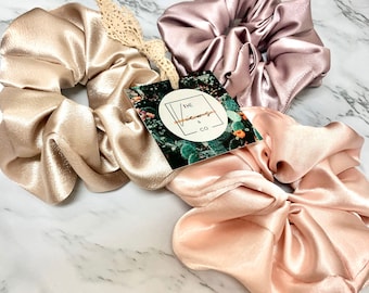May include: Three satin scrunchies in shades of blush, light pink, and gold. The scrunchies are arranged on a white marble surface. A small tag with the text "THE VIENNS CO" is visible in the center of the image.