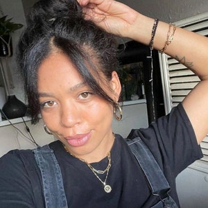 May include: A woman with dark hair in a bun wears a black t-shirt with a gold chain necklace and a gold pendant. She is wearing hoop earrings and has a tattoo on her arm.