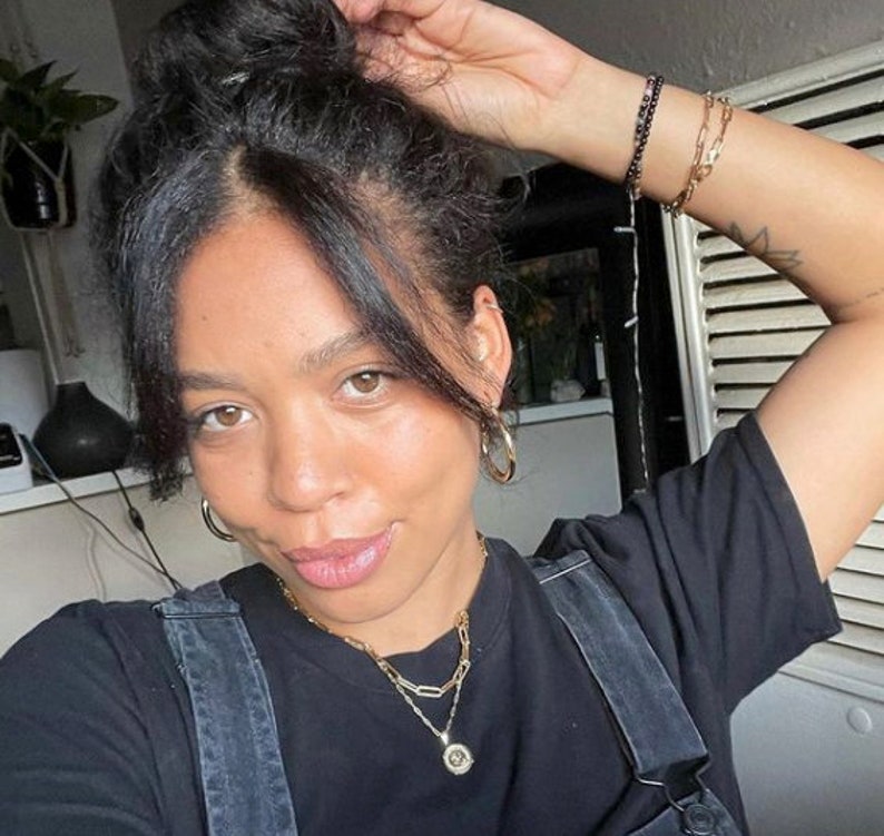 May include: A woman with dark hair in a bun wears a black t-shirt with a gold chain necklace and a gold pendant. She is wearing hoop earrings and has a tattoo on her arm.