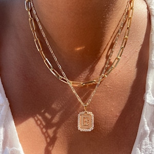 Może przedstawiać: Złoty naszyjnik z łańcuszkiem i kwadratowym wisiorem z pojedynczą literą 'B' otoczoną małymi, przezroczystymi kamieniami.