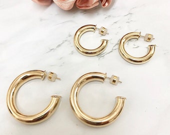May include: Four pairs of gold-colored hoop earrings in varying sizes. The earrings are made of a shiny, smooth metal and have a classic, rounded design. The earrings are displayed on a white marble surface, with a pink scrunchie in the background.