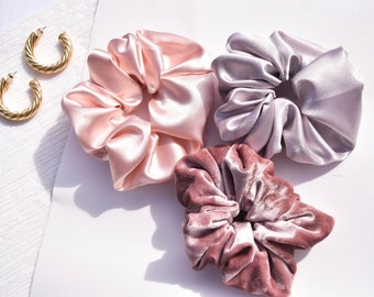 May include: Three hair scrunchies in different colors and textures. One is a light pink satin scrunchie, another is a light gray satin scrunchie, and the last is a dark pink velvet scrunchie.