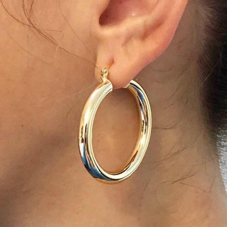May include: Gold hoop earrings with a thick, rounded band. The earrings are large and have a simple, classic design.