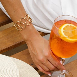 May include: A gold-toned, open-ended, wavy bracelet on a person's wrist. The person is holding a glass of orange juice with a slice of orange in it.