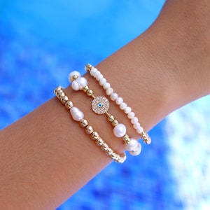 Puede incluir: Tres pulseras en una muñeca. La pulsera superior es blanca con pequeñas perlas. La pulsera del medio es dorada con pequeñas cuentas. La pulsera inferior es blanca con perlas más grandes. Un dije dorado con un diseño de ojo azul maligno cuelga de la pulsera del medio.