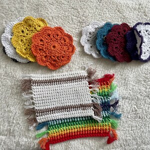 Peut inclure: Un ensemble de dessous-de-verre en crochet de différentes couleurs, dont le jaune, l'orange, le blanc, le bleu sarcelle, le bordeaux et le bleu marine. Les dessous-de-verre sont disposés en deux groupes, avec un dessous-de-plat en crochet blanc à rayures brunes et un dessous-de-plat en crochet arc-en-ciel avec des franges en bas.