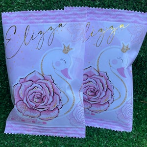May include: Two pink and white favor bags with a gold glitter design. The bags feature a white swan with a gold crown, a pink rose, and the name "Elizza" in gold script.