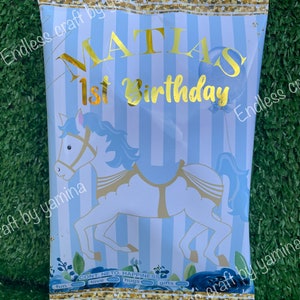 May include: A party favor bag with a blue and white striped background. The bag features a carousel horse illustration and gold text that reads "MATIAS 1st Birthday." The top and bottom edges are trimmed with gold foil.