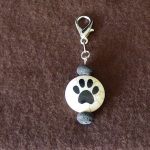 May include: A silver charm with a black paw print on a white round bead, with two black lava rock beads on either side of the paw print bead. The charm is attached to a silver lobster clasp.