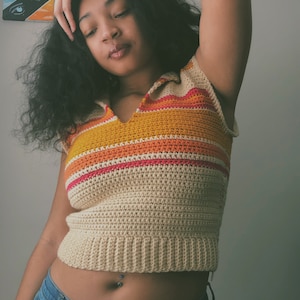 May include: A sleeveless crochet polo shirt featuring a beige base with horizontal stripes in vibrant orange, yellow, and pink. The garment has a collared neckline, a V-shaped front opening, and a ribbed hemline, paired with blue denim jeans.