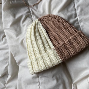May include: A crocheted beanie hat with a white and brown color block design. The hat is made with a soft, textured yarn.
