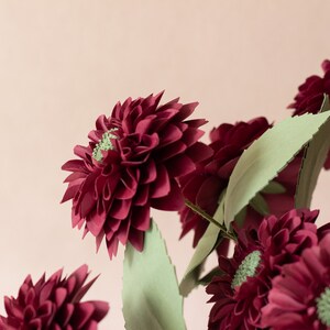 May include: A bouquet of burgundy paper flowers with green paper leaves. The flowers are arranged in a vase, and the background is a light pink.