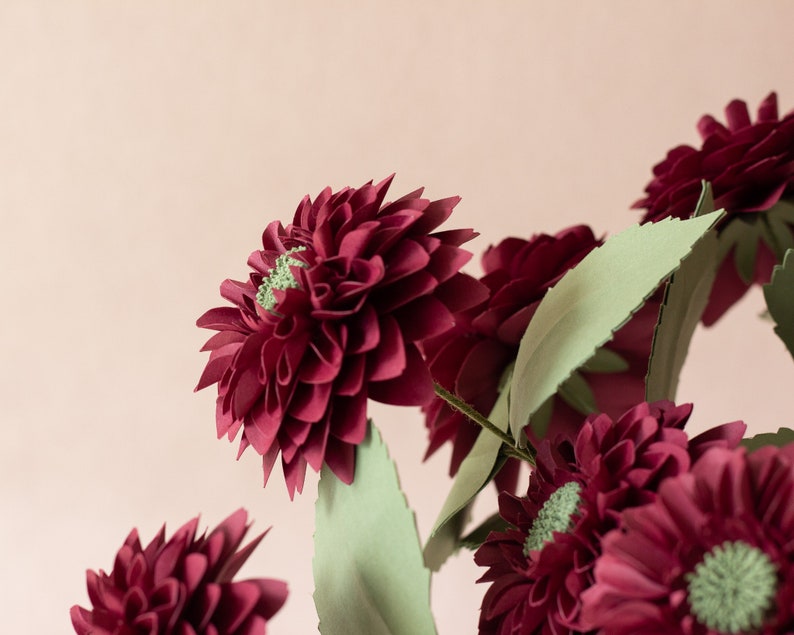 May include: A bouquet of burgundy paper flowers with green paper leaves. The flowers are arranged in a vase, and the background is a light pink.