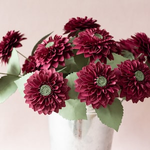 May include: A bouquet of burgundy paper dahlias in a silver bucket. The flowers are arranged in a cluster with green paper leaves.