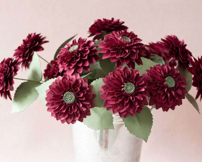 May include: A bouquet of burgundy paper dahlias in a silver bucket. The flowers are arranged in a cluster with green paper leaves.