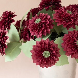 May include: A bouquet of burgundy paper dahlias in a white ceramic vase. The flowers are arranged in a cluster with green paper leaves.