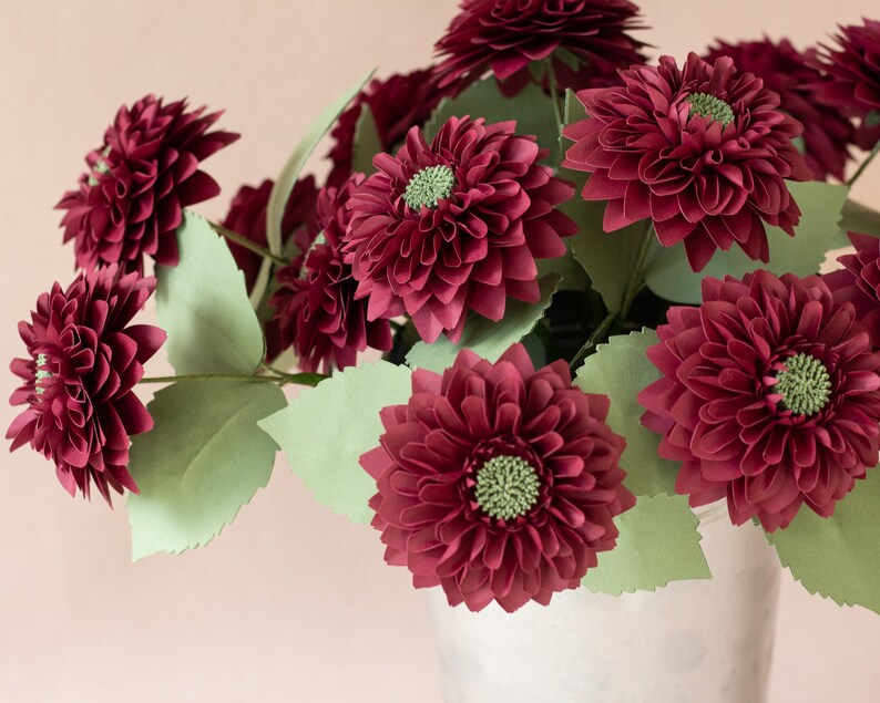 May include: A bouquet of burgundy paper dahlias in a white ceramic vase. The flowers are arranged in a cluster with green paper leaves.