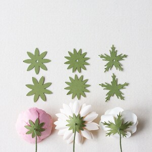 May include: Three paper flowers with green paper leaves attached to the back. The flowers are pink, white, and white. The flowers are arranged in a row on a white background.