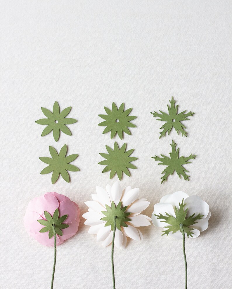 May include: Three paper flowers with green paper leaves attached to the back. The flowers are pink, white, and white. The flowers are arranged in a row on a white background.