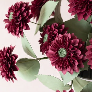 May include: A bouquet of burgundy paper dahlias with green paper leaves. The flowers are arranged in a vase.