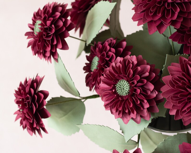 May include: A bouquet of burgundy paper dahlias with green paper leaves. The flowers are arranged in a vase.