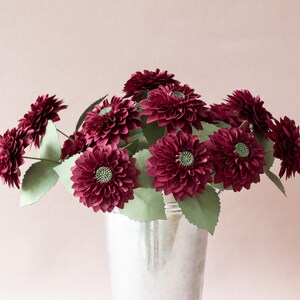 May include: A bouquet of burgundy paper dahlias in a silver vase. The flowers are arranged in a loose, natural style, with the stems extending out from the vase.