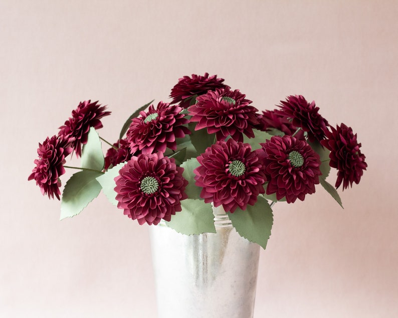 May include: A bouquet of burgundy paper dahlias in a silver vase. The flowers are arranged in a loose, natural style, with the stems extending out from the vase.