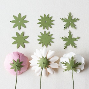 May include: Three paper flowers with green paper leaves attached to the back. The flowers are pink, white, and white. There are six green paper leaves on the table.