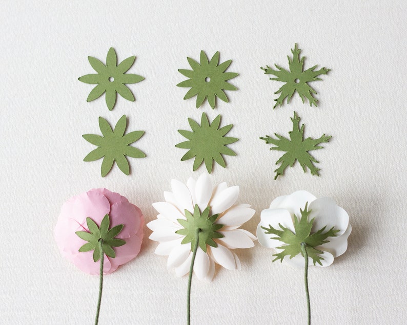 May include: Three paper flowers with green paper leaves attached to the back. The flowers are pink, white, and white. There are six green paper leaves on the table.