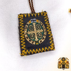 K&ouml;nnte beinhalten: Ein kleiner, dunkelbrauner Stoffanh&auml;nger mit einem goldenen und gr&uuml;nen Stickmuster. Der Anh&auml;nger ist von einem gelben Zickzack-Stich umgeben und hat eine braune Schnur.