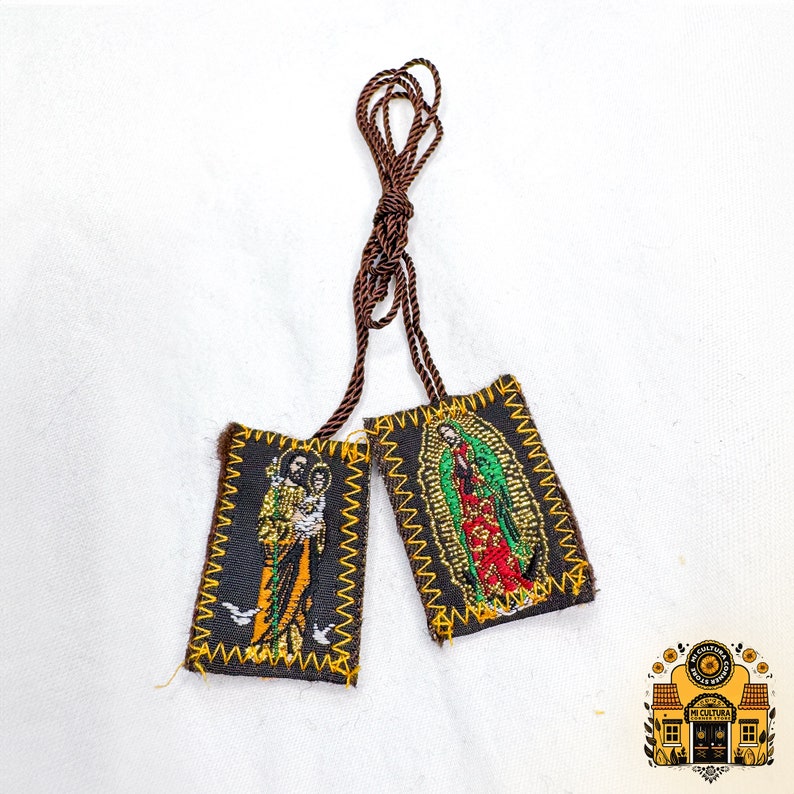 Pode incluir: Dois pequenos amuletos de tecido castanho com guarni&ccedil;&otilde;es douradas e imagens bordadas de figuras religiosas. Os amuletos est&atilde;o presos a um cord&atilde;o castanho.
