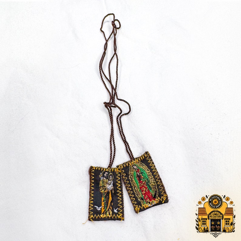 Pode incluir: Dois pequenos amuletos de tecido retangulares bordados com imagens de figuras religiosas. Os amuletos est&atilde;o presos a um cord&atilde;o castanho.