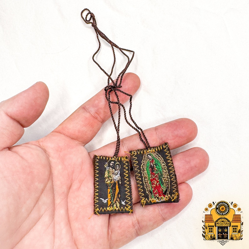 Pode incluir: Dois pequenos amuletos religiosos de tecido castanho com bordados dourados. Um amuleto apresenta uma imagem da Virgem Maria, o outro apresenta uma imagem de Jesus e uma pomba. Os amuletos est&atilde;o presos a um cord&atilde;o castanho.