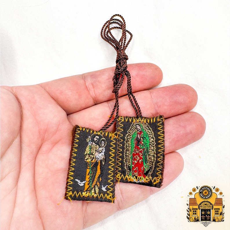 Pode incluir: Dois pequenos amuletos de ora&ccedil;&atilde;o de tecido castanho com bordados dourados. Um amuleto apresenta uma imagem de S&atilde;o Jos&eacute; segurando o menino Jesus. O outro amuleto apresenta uma imagem de Nossa Senhora de Guadalupe. Os amuletos est&atilde;o unidos a um cord&atilde;o castanho.
