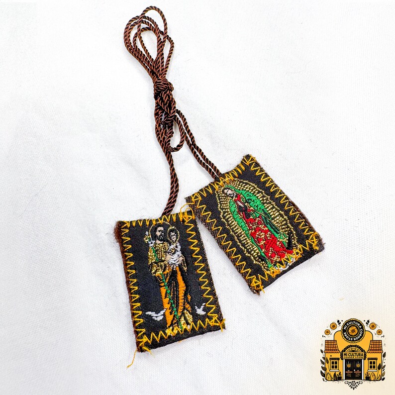 Pode incluir: Dois pequenos amuletos de tecido retangulares bordados com imagens de figuras religiosas. Os amuletos est&atilde;o presos a um cord&atilde;o castanho. A imagem no amuleto da esquerda &eacute; de um homem que segura um beb&ecirc;. A imagem no amuleto da direita &eacute; de uma mulher com uma t&uacute;nica azul.
