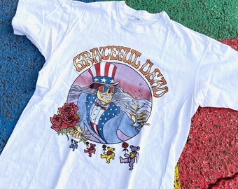 Puede incluir: Camiseta blanca con un gráfico de Tío Sam, un esqueleto con un sombrero de copa y sosteniendo un cóctel, con el texto "Grateful Dead" alrededor de la imagen. Hay cinco osos bailando debajo de la imagen.