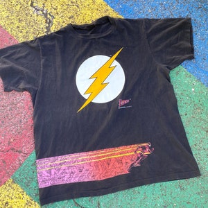 Puede incluir: Camiseta negra con un círculo blanco y un rayo amarillo. El texto "The Flash" está impreso debajo del gráfico. La parte inferior de la camiseta tiene un gráfico de Flash corriendo.