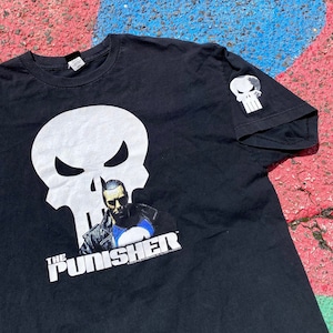 May include: Black t-shirt with a white skull graphic and the text "The Punisher" in white. The skull has a menacing expression and the character is wearing a black leather jacket. There is a smaller white skull graphic on the right sleeve.