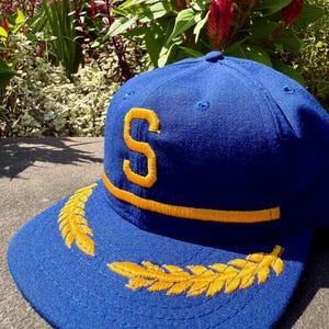 May include: A royal blue baseball cap featuring a prominent yellow embroidered letter S on the front panel. The cap includes a horizontal yellow embroidered line above the brim and two yellow embroidered laurel leaf designs on the brim.