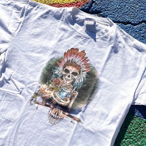 May include: White t-shirt with a graphic print of a skeleton wearing a Native American headdress. The skeleton is holding a staff and a skull. The graphic is in shades of green, red, blue, and yellow.
